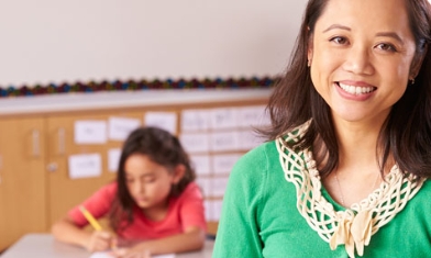 Smiling female teacher in green cardigan in foreground, child student writing in background