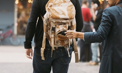 Man with rolled sleeve taking item from backpack on another man walking in ahead in a street