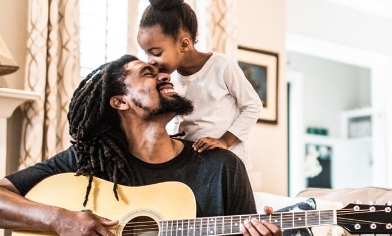 Little girl kissing her daddy on the forehead while he's playing a guitar, in their home