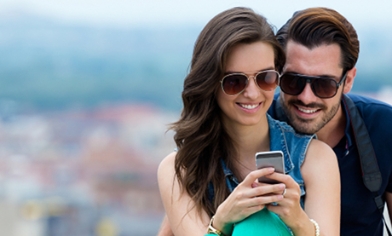 A couple wearing sunglasses and smiling while looking at something on the woman's phone in her hands