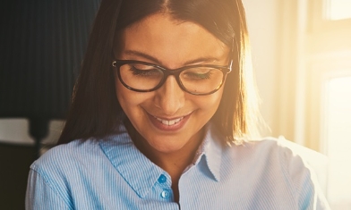 Young smiling woman in light blue shirt wearing glasses, sat in a sunlit lounge, looking at laptop