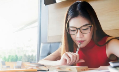 Woman sat in a well-lit café, holding pen to her mouth while reading from a book for her studies