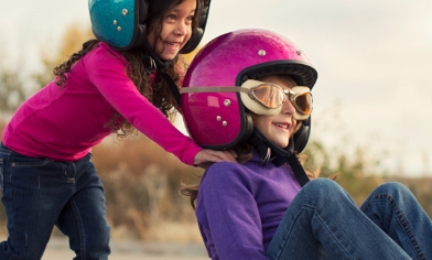 Two smiling girls wearing helmets, one sat on skateboard wearing goggles, being pushed by the other