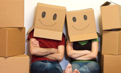 Couple sat between stacked boxes, both wearing boxes on their heads with smiley faces drawn on them