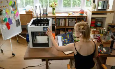 A person seated at a desk in a craft room, operating a 3D printer.