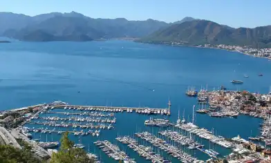 a marina with rows of boats and yachts docked along a calm coastline, surrounded by mountainous terrain and nearby buildings