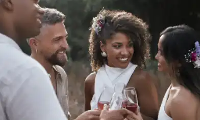People dressed in light-coloured outfits clinking glasses of wine outdoors, with floral hair accessories visible