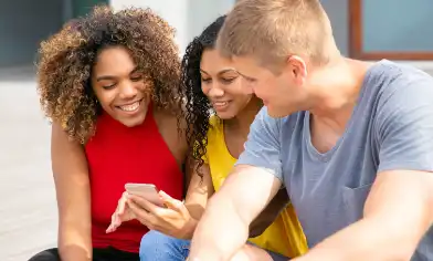 Three young friends in bright clothes smile at something on one of their phones while sitting on the steps of their university building.