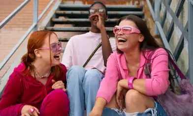 Three teens sit on outdoor stairs with brick walls and railings behind them. They wear red, white and pink outfits and sunglasses