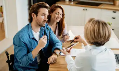 Three people at a kitchen table, one in a wheelchair holding a pen, discussing documents. One holds out a digital tablet for the others to read