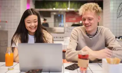Two smiling people are hanging out at a café table with snacks and drinks, looking at a laptop. The surrounding walls and tabletop are made of white tiles