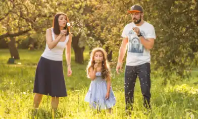 A family stands in a sunlit meadow blowing dandelion seeds surrounded by tall grass and trees