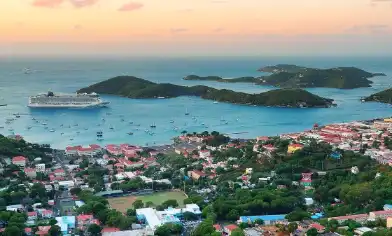 Aerial view of a coastal town with a large cruise ship docked near small islands, surrounded by buildings, roads, green spaces and boats, under an orange and pink sky 