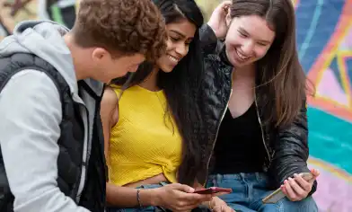 Three teens sit in front of a bright graffiti wall, checking something out on a phone together 