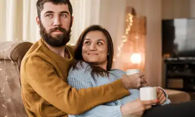 A couple sit at home, arms around each other and holding coffee cups