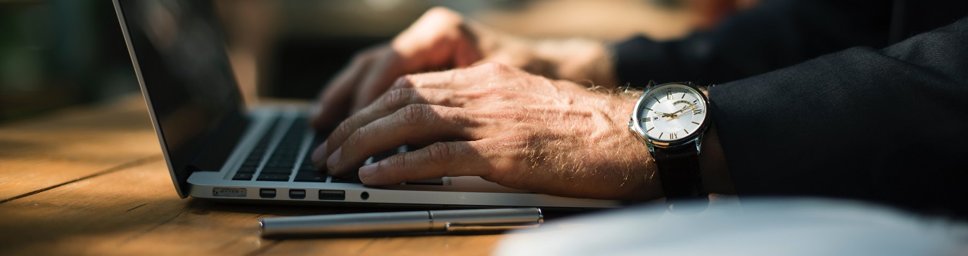 A man's hand, wearing a watch, typing on his laptop on his desk