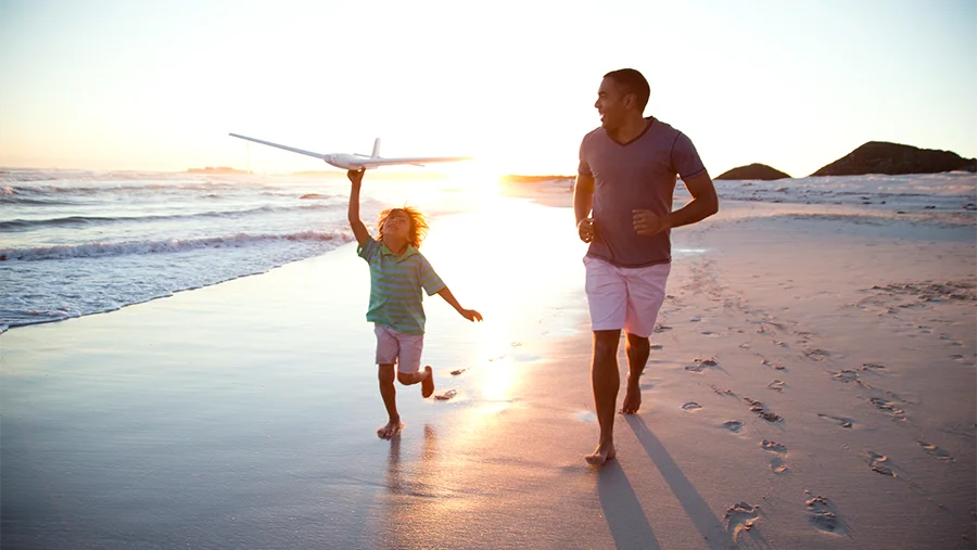Father and son who is holding a model plane above his head running along a wet sun set soaked beach