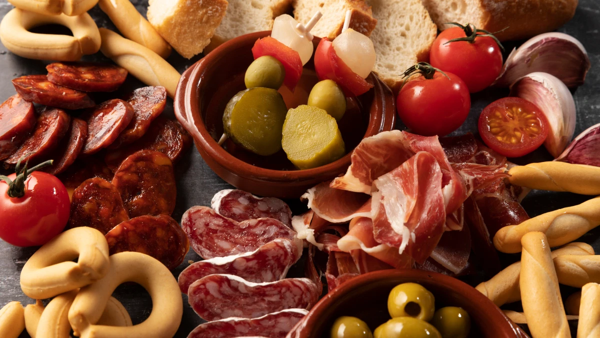 A spread of appetisers including cured meats, olives, pickles, cherry tomatoes, bread, garlic and breadsticks arranged on a surface