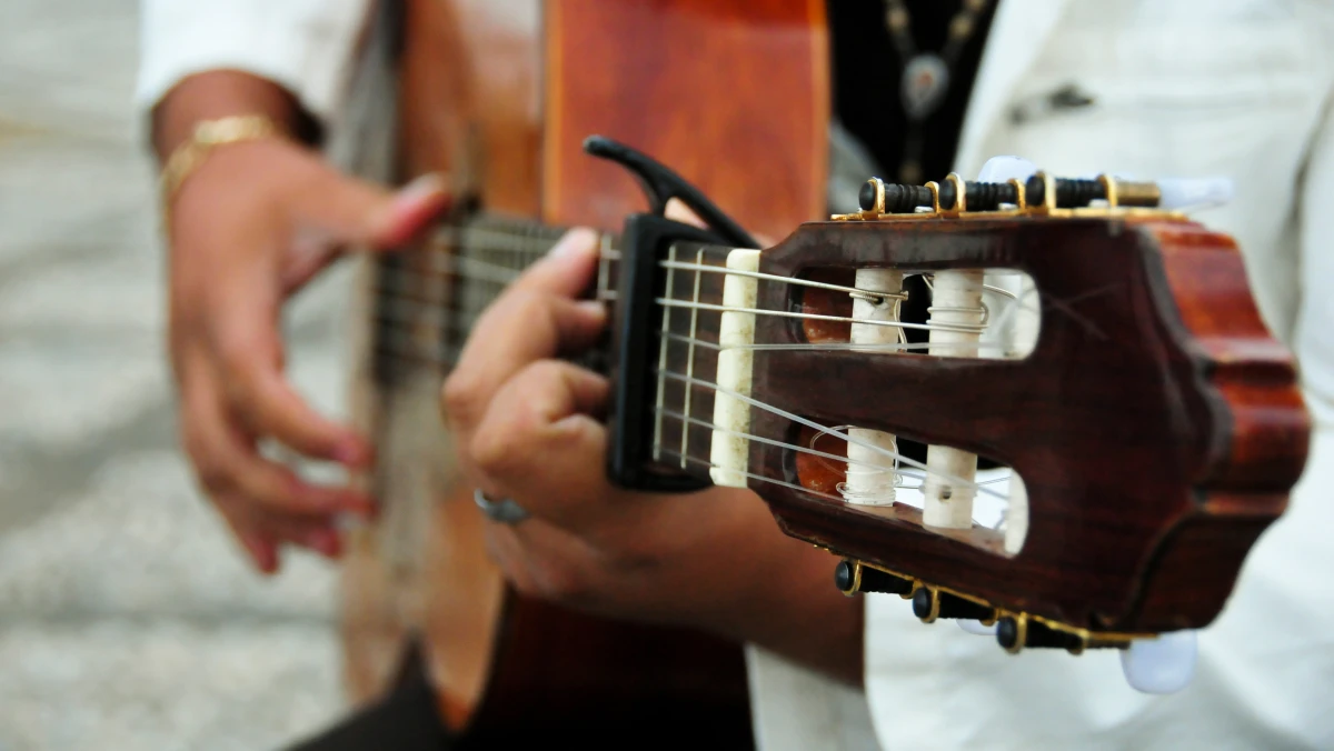 A close-up of someone playing an acoustic guitar, with one hand strumming and the other on the fretboard, wearing a white shirt and beaded necklace