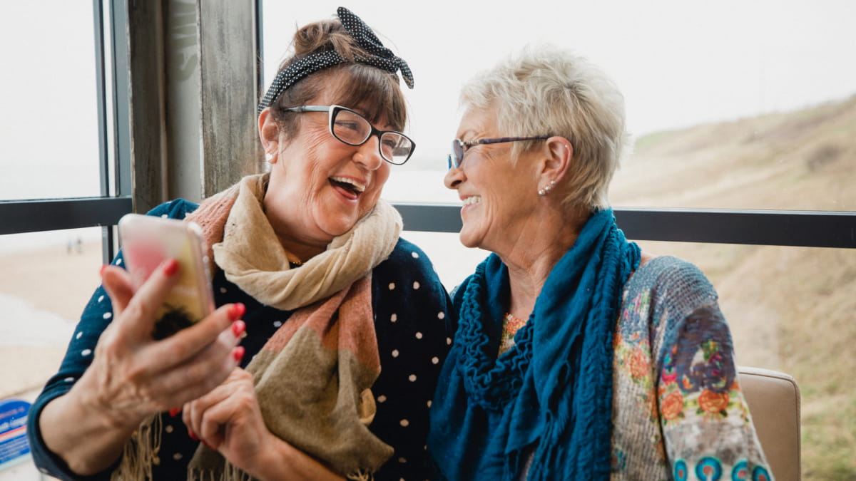 Two mature ladies looking at each other laughing, one with a phone in her hand held up high