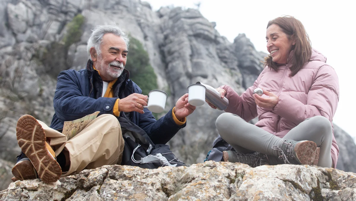 Older man and woman smiling and toasting with mugs while sitting on a rocky mountain ledge