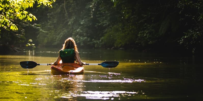 Woman wearing green backpack in kayak holding paddle, floating in green waters surrounded by greenery