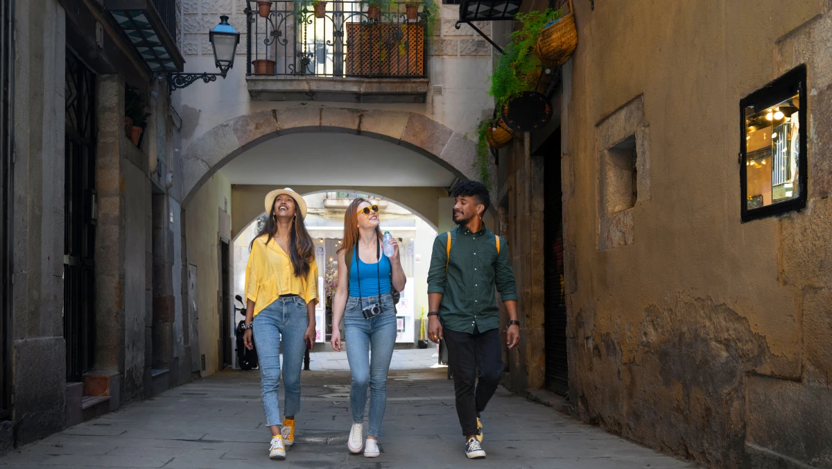 Three people walk down a narrow street with an archway, flanked by buildings and hanging plants, with a balcony above