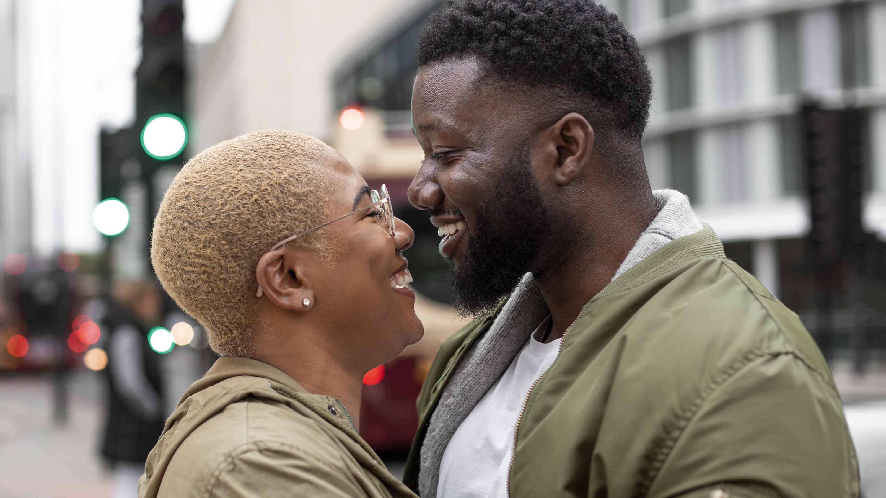 Two people stand close together on a city street, both wearing green jackets, with traffic lights and buildings in the background