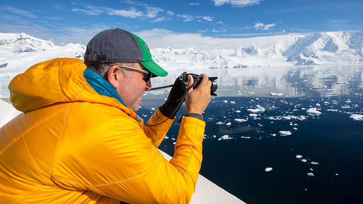Man in an arctic scene wearing a yellow winter and cap, holding a camera up taking a picture