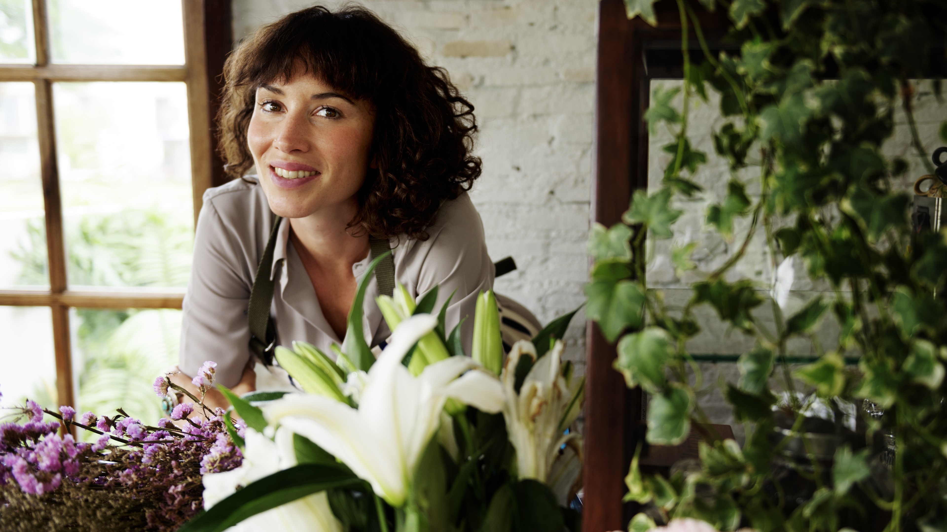 A person with curly hair stands behind an arrangement of white lilies and pink roses, surrounded by green plants in a naturally lit indoor setting 