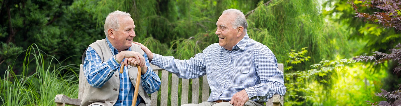 Two elderly men sat on a park bench, one with his hand on others shoulder, smiling at each other