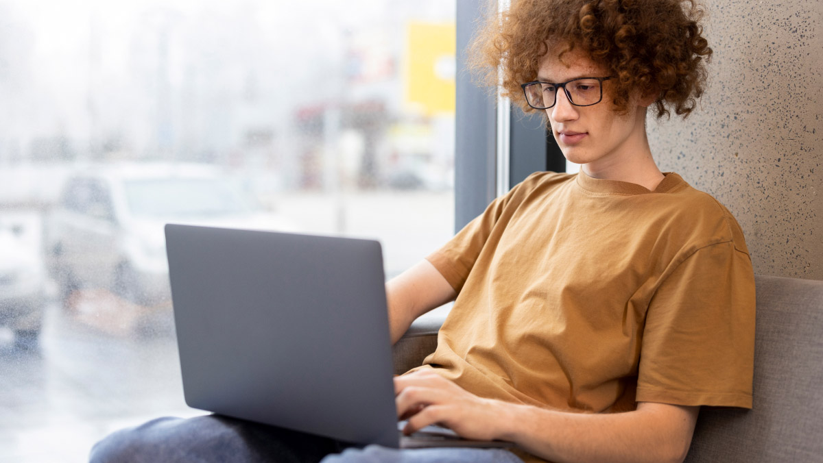 Someone in a brown T-shirt and jeans is sitting on a couch using a laptop, with large windows behind them showing buildings and cars outside