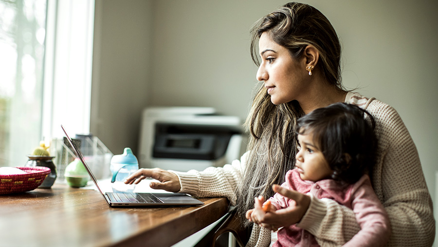 Young mother sat at a table using her laptop and holding her little girl on her lap