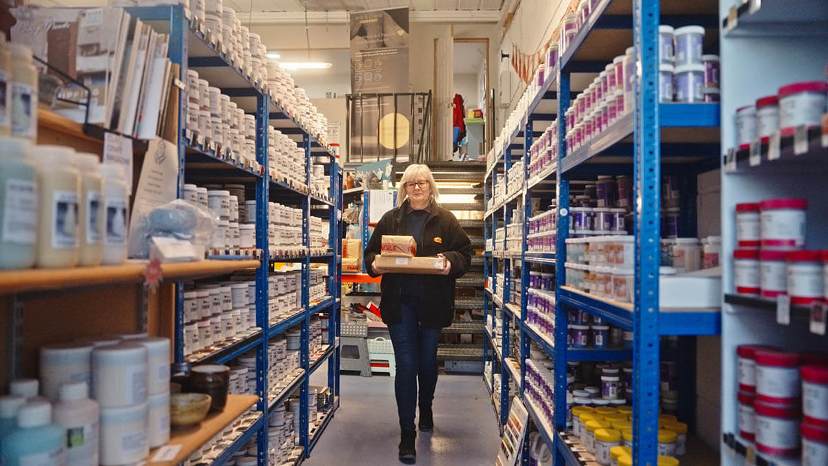 A person walking down an aisle in a store or warehouse, carrying several items, with shelves stocked with containers and bottles on both sides