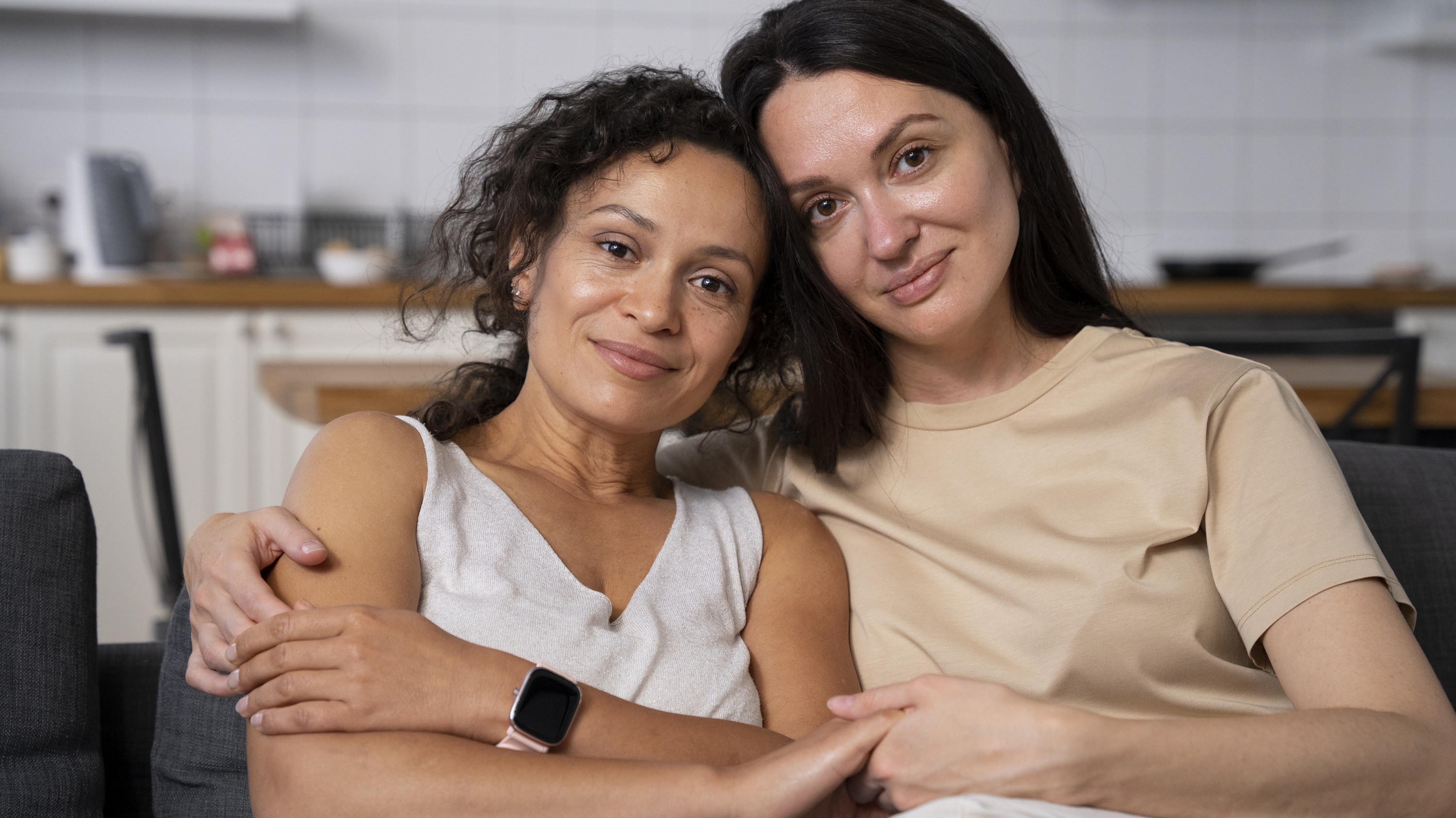 Two people sit together on a couch, their heads leaning together and smiling, with kitchen items in the background
