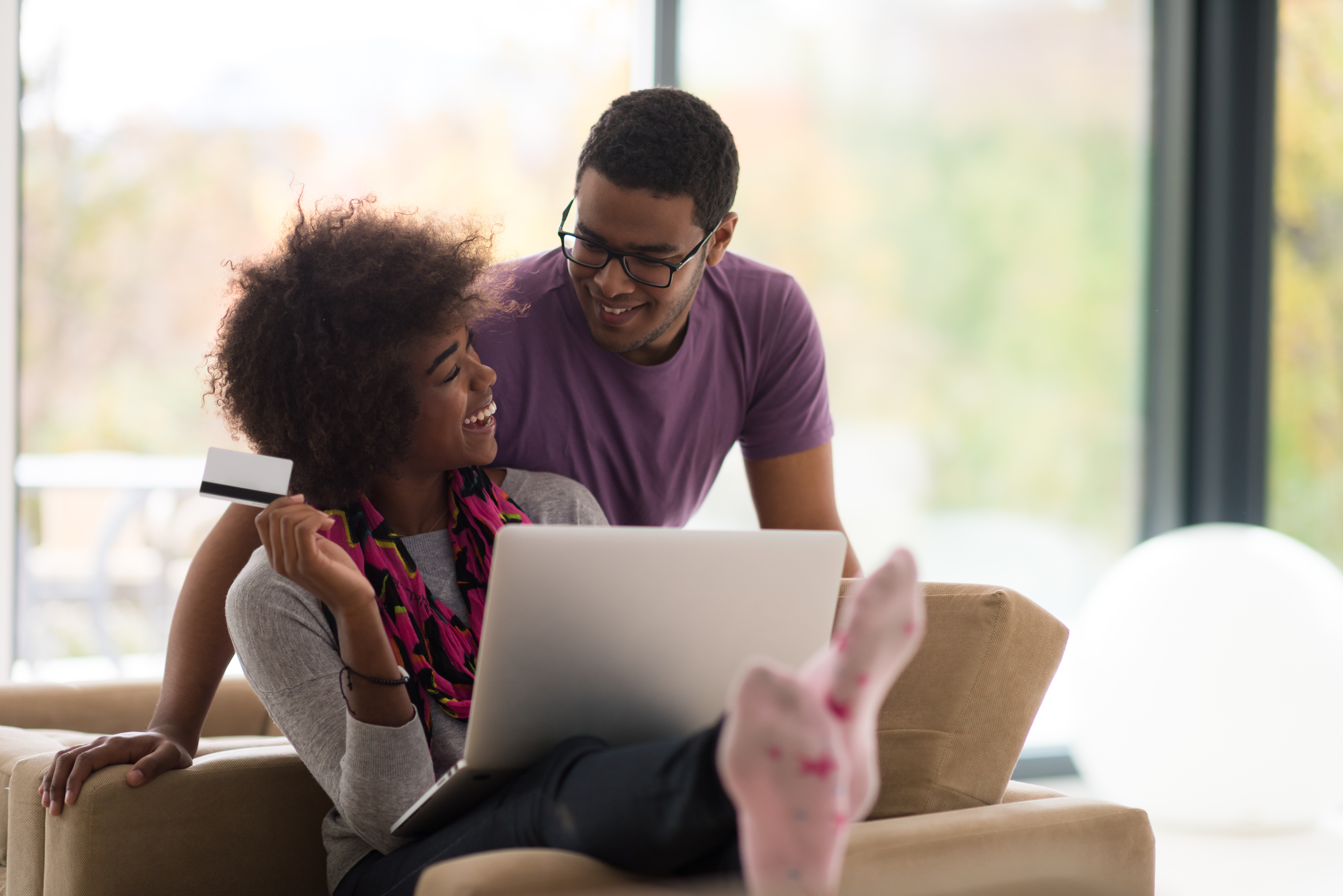 Woman and partner laughing together, whilst she is sitting down with laptop, holding a credit card in one hand