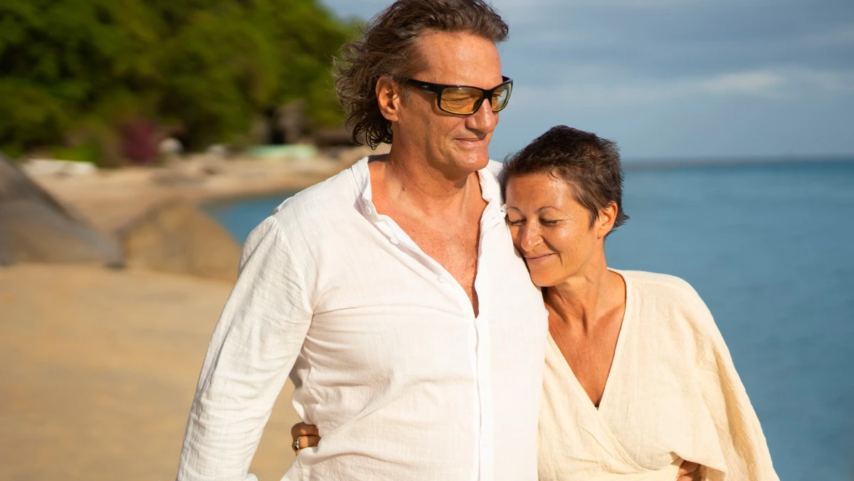Two people stand on a sandy beach near large rocks and greenery, with the ocean and a partly cloudy sky in the background