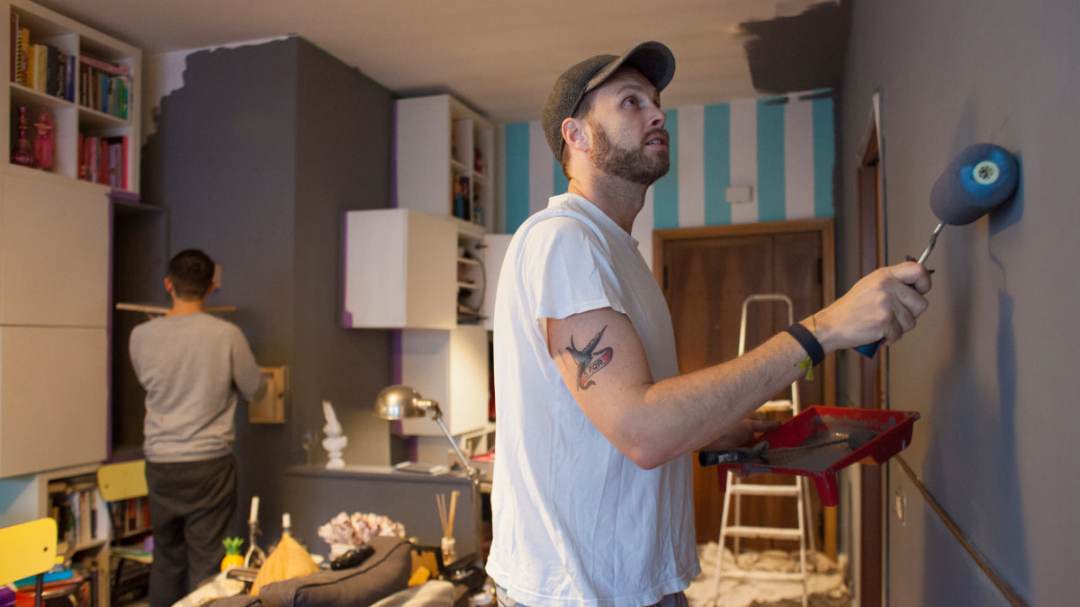 Two men painting a room, man on left with his back to us, other on the right using a paint roller