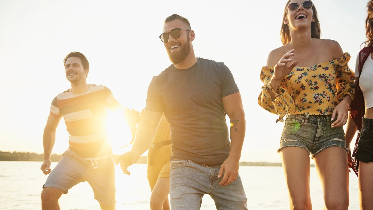 A group of people stand near a body of water at sunset or sunrise, dressed in casual summer clothes, with warm light in the background