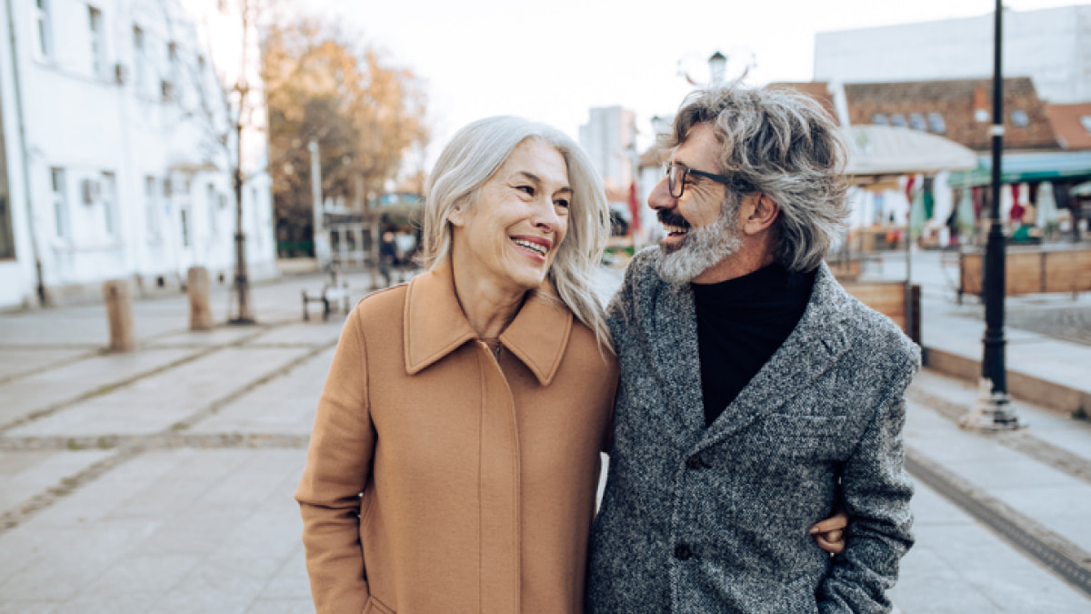 Older couple in wool coats walking down a street smiling and looking at one another