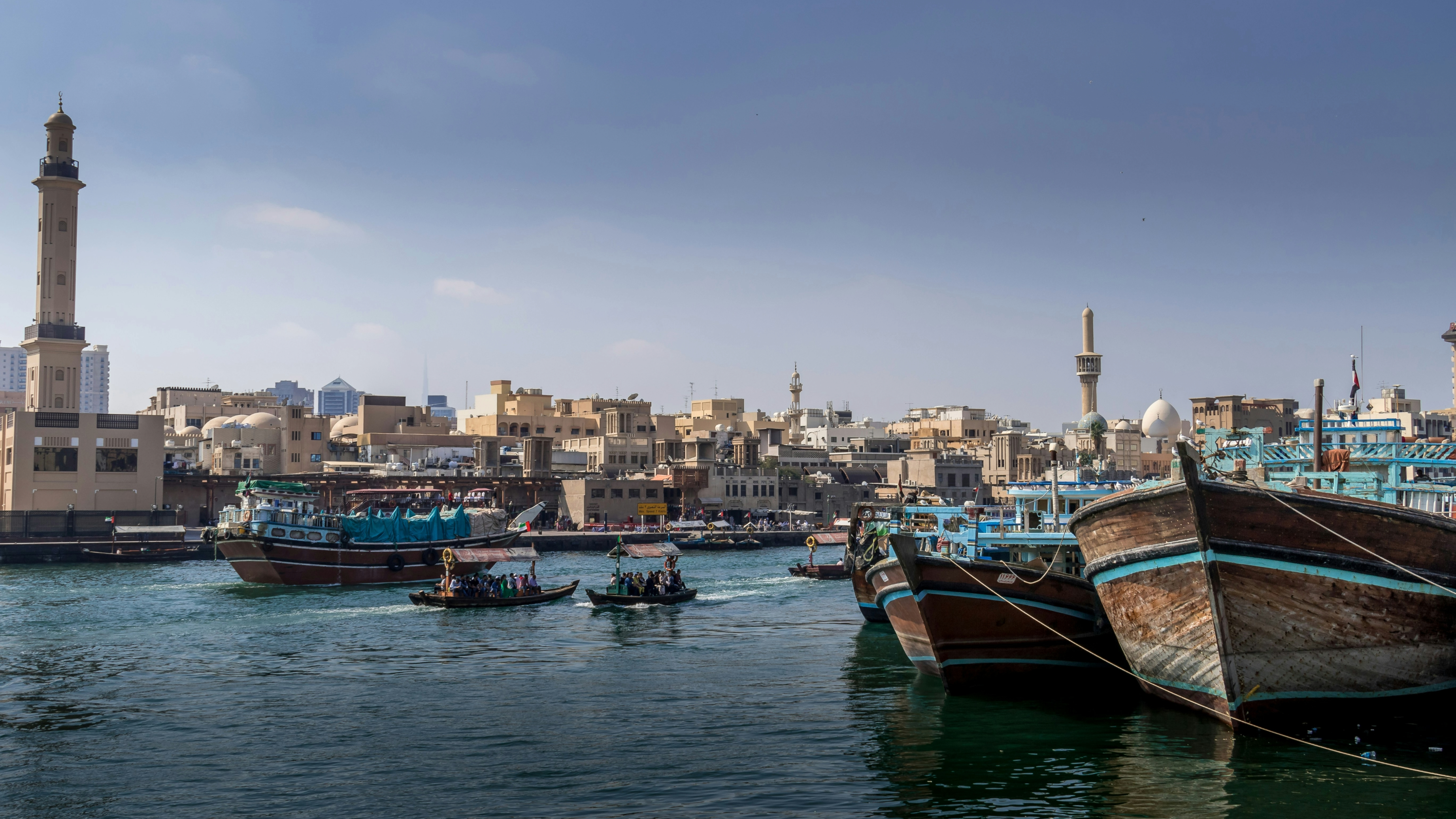 Traditional wooden boats on the water at Dubai Marina, with low-rise buildings and a mosque minaret in the background