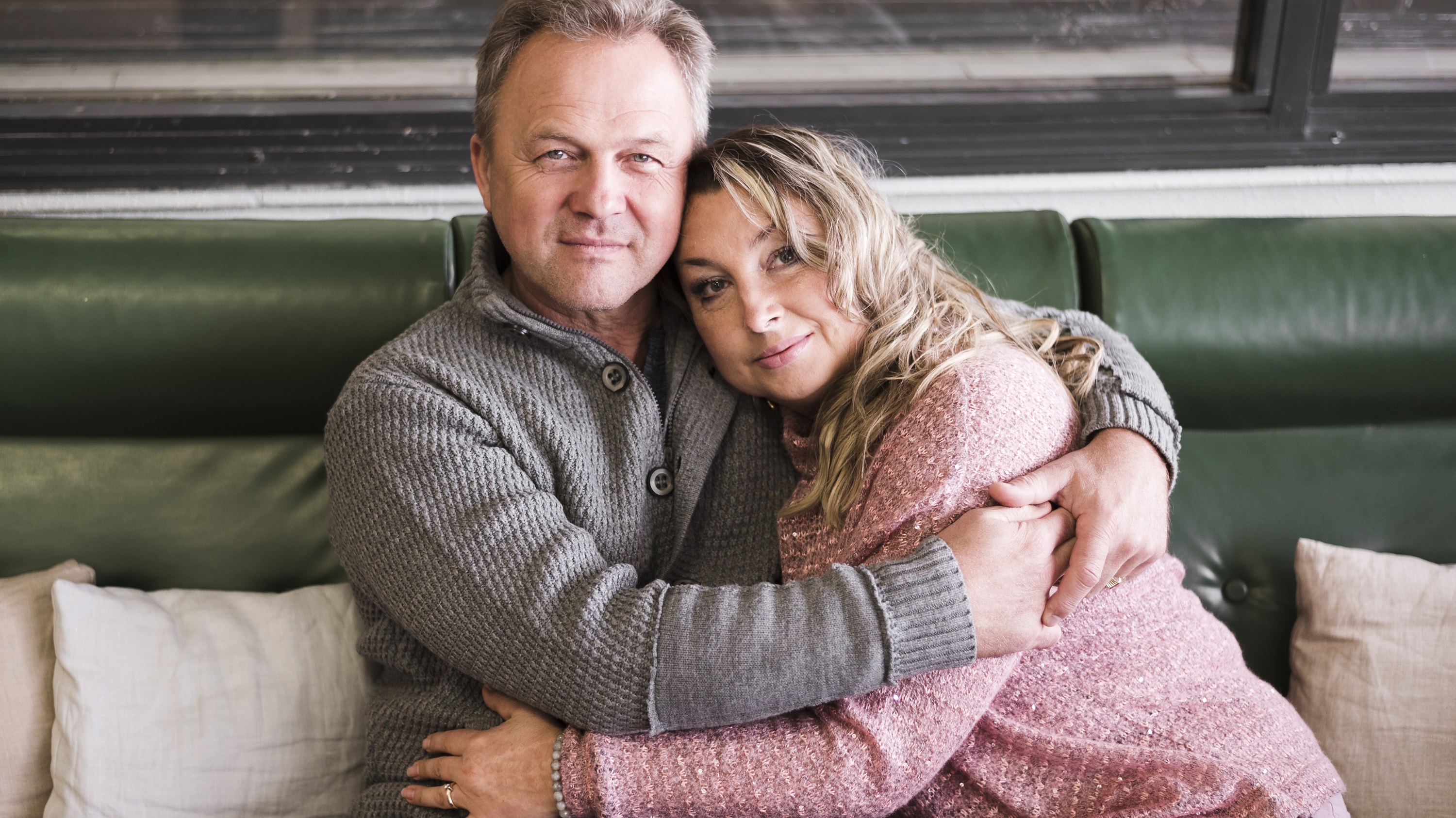 Two people sit closely on a green couch with arms around each other, surrounded by cushions