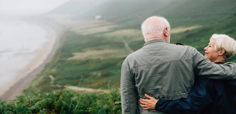 Elderly couple looking at each other lovingly, stood on a high hillside overlooking coastline below