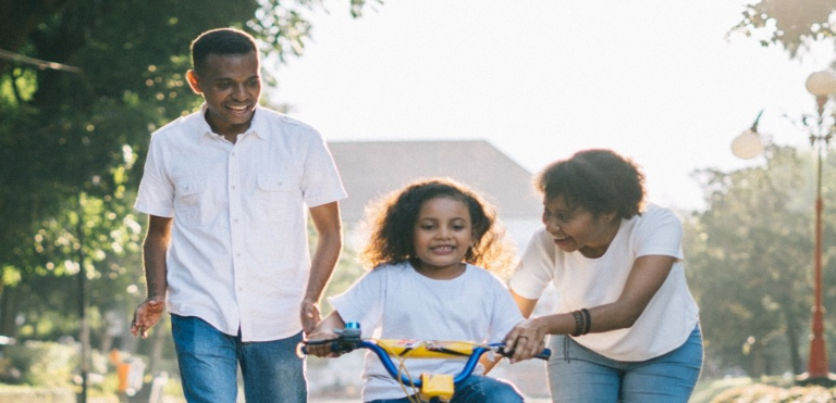 Mother and father teaching their little girl to ride a bike along a sunlit road