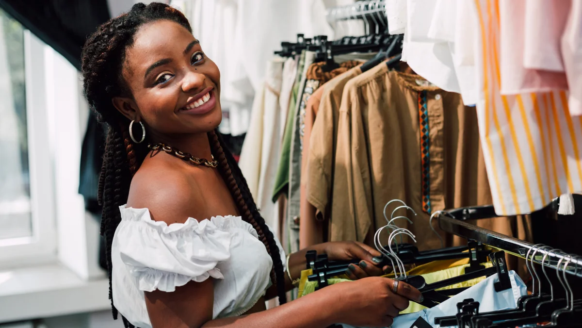 A smiling person wearing a white top sorts clothes on hangers in a clothing store, with various shirts and jackets visible 