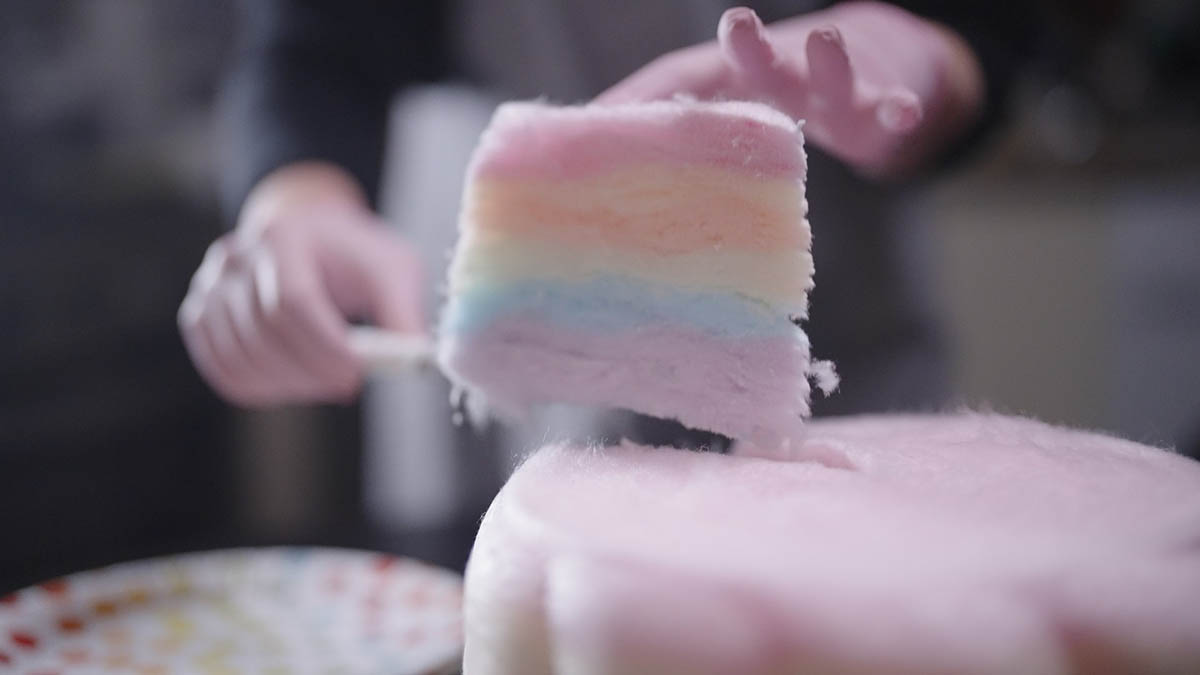A close-up of a person lifting a slice of pastel-coloured candy floss cake with a utensil, showing pink, yellow, blue and purple layers 