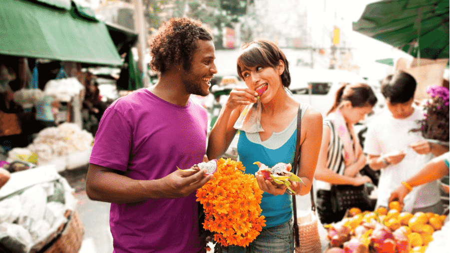 A man and woman in a street market are looking at each other, both smiling, as the woman eats fruit