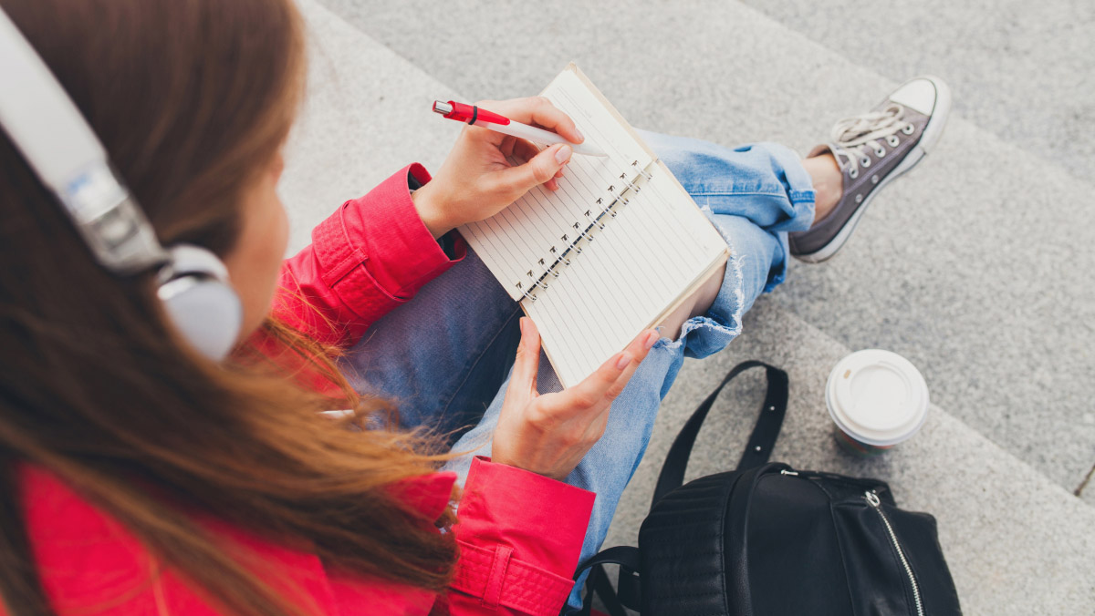 A person in a red jacket and jeans sits on steps with headphones on, writing in a notebook next to a backpack and coffee cup