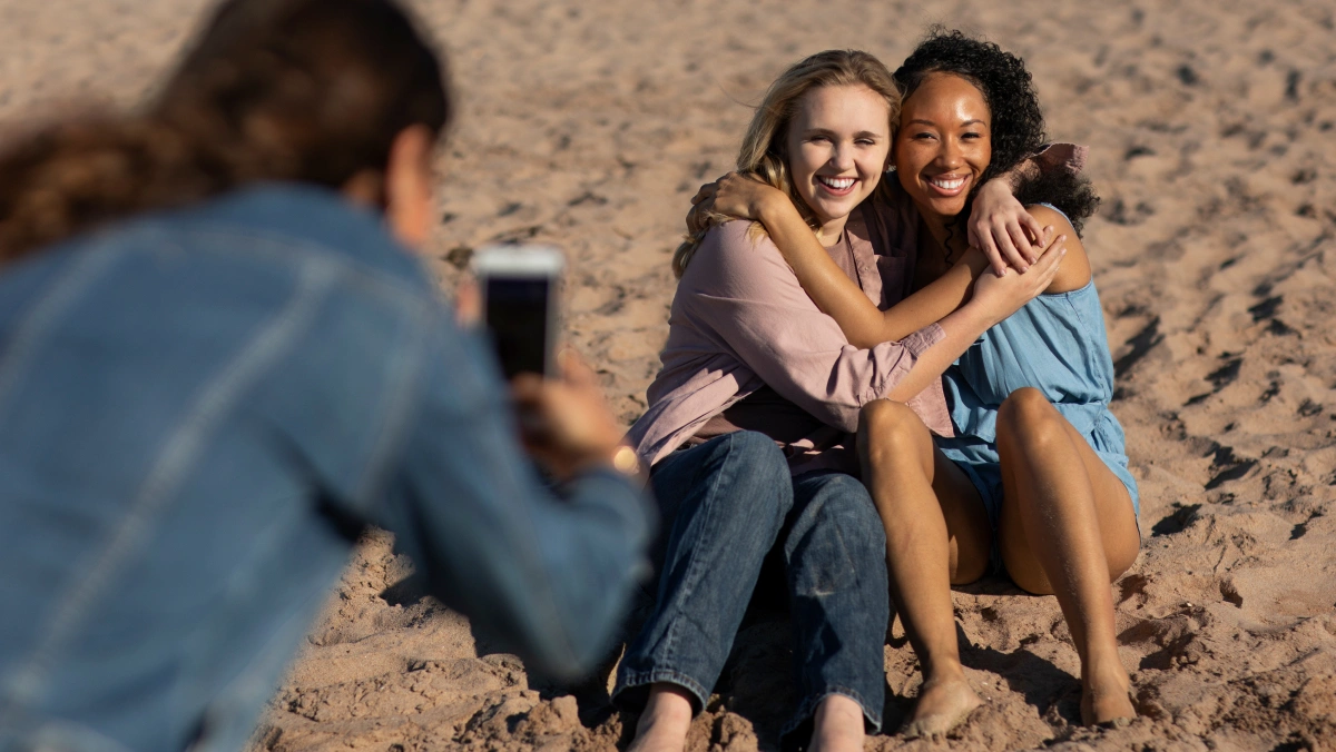 a person with a smart phone taking a picture of two friends hugging on a beach