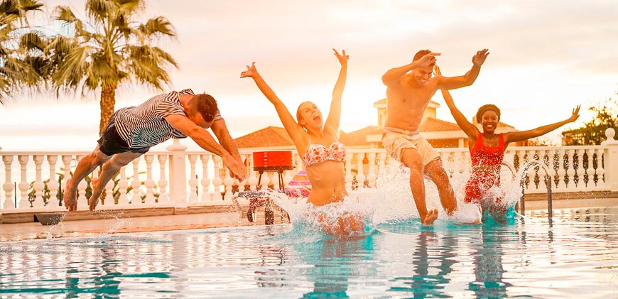 Two young men and two young women jumping in a pool with trees, houses and beach in distance